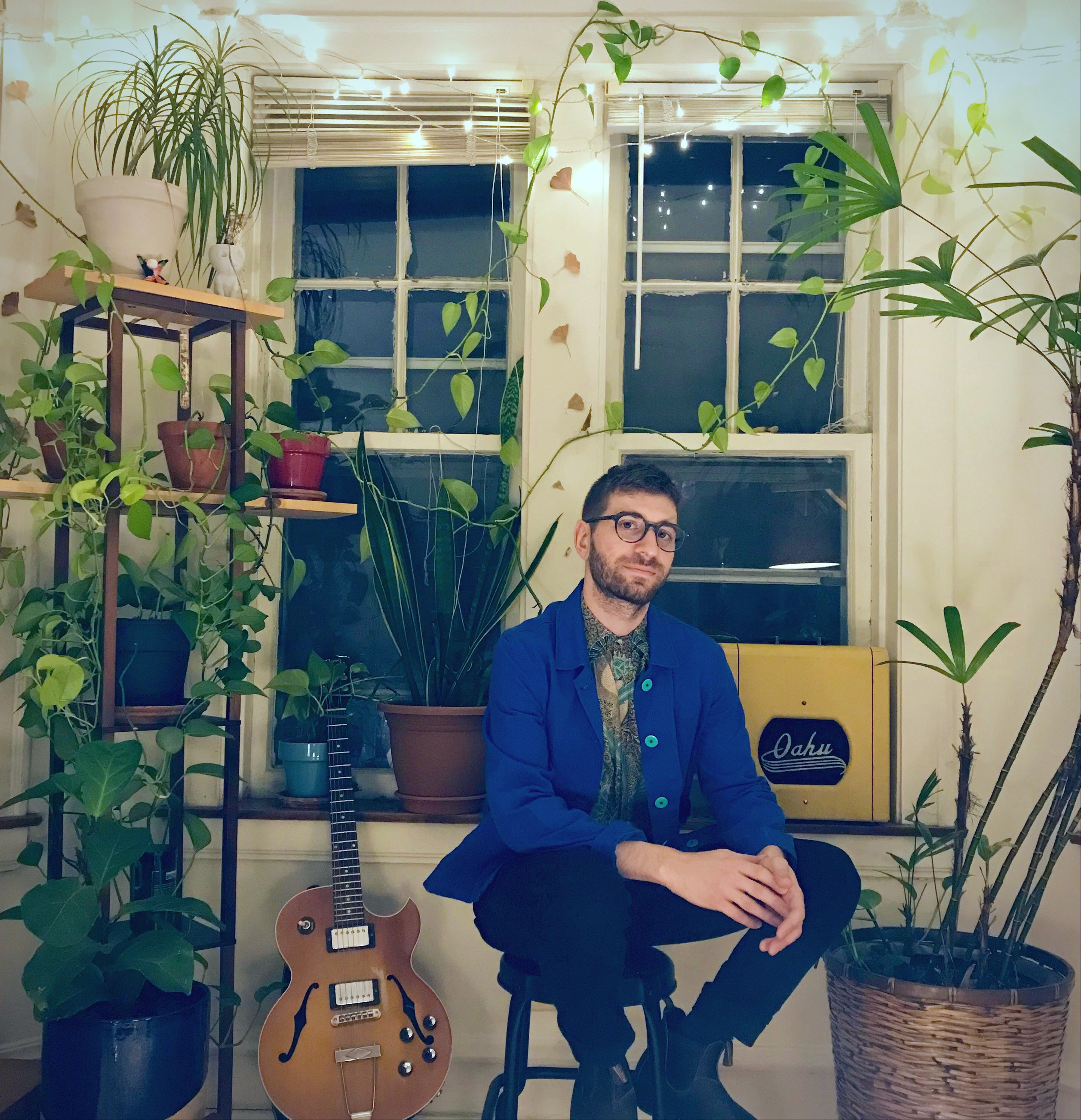 Matt Gold sitting on a stool in front of a window, surrounded by tall green plants Matt Gold sitting on a stool in front of a window, surrounded by tall green plants