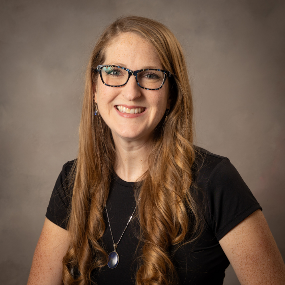 Smiling woman with long blonde hair and glasses, wearing a necklace and a black top, in front of a solid grey background. Smiling woman with long blonde hair and glasses, wearing a necklace and a black top, in front of a solid grey background.