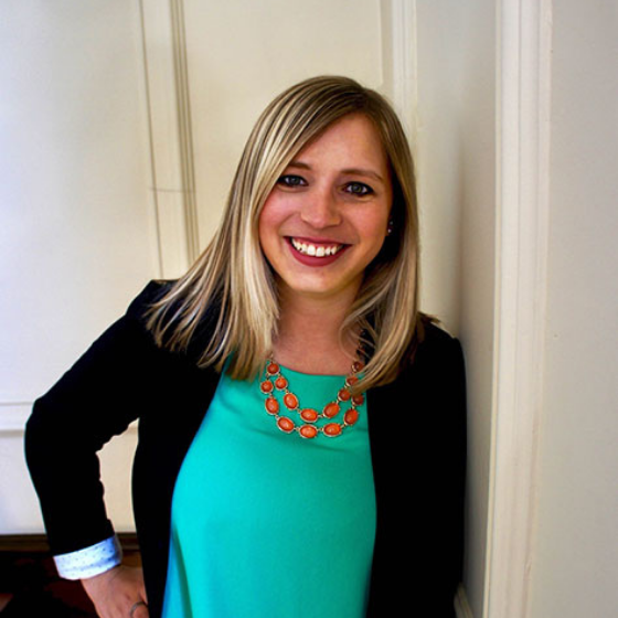 Smiling woman with straight blonde hair, wearing a teal top, black blazer, and an orange necklace, leaning against an off-white colored wall. Smiling woman with straight blonde hair, wearing a teal top, black blazer, and an orange necklace, leaning against an off-white colored wall.