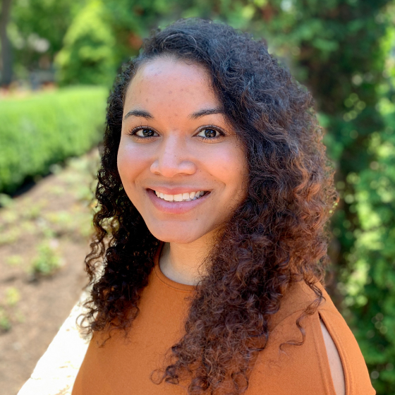 Smiling woman with curly, mid-length hair and an orange top standing outdoors, with greenery in the background. Smiling woman with curly, mid-length hair and an orange top standing outdoors, with greenery in the background.