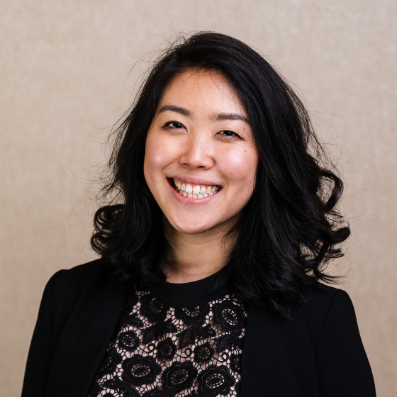 Smiling woman with wavy, mid-length black hair, wearing a black lace top with a black blazer, against a solid beige background. Smiling woman with wavy, mid-length black hair, wearing a black lace top with a black blazer, against a solid beige background.