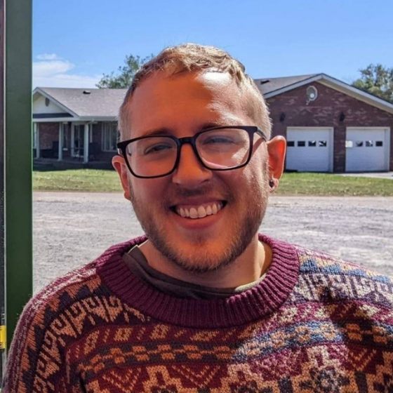 Smiling man with glasses, wearing a knit sweater, pictured in front of the backyard of a house. Smiling man with glasses, wearing a knit sweater, pictured in front of the backyard of a house.