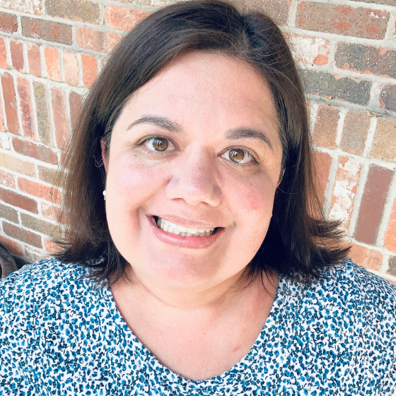 Smiling woman with mid-length brown hair, wearing a floral blue top, leaning against a brick wall. Smiling woman with mid-length brown hair, wearing a floral blue top, leaning against a brick wall.