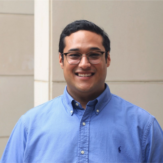 Smiling man with short dark brown hair, wearing glasses and a blue long sleeved top with a collar, against a white wall. Smiling man with short dark brown hair, wearing glasses and a blue long sleeved top with a collar, against a white wall.