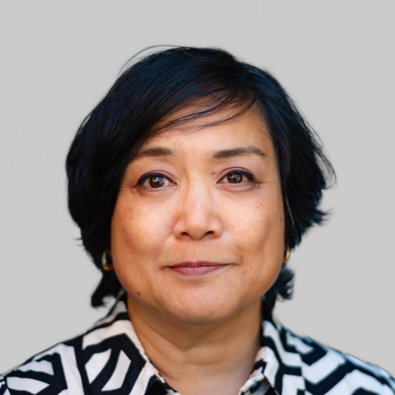 Woman with short black hair, wearing a black and white striped blouse, pictured against a solid grey background. Woman with short black hair, wearing a black and white striped blouse, pictured against a solid grey background.