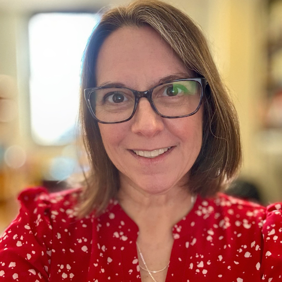 Smiling woman with shoulder length brown hair, wearing glasses and a red top, against a blurred room background. Smiling woman with shoulder length brown hair, wearing glasses and a red top, against a blurred room background.