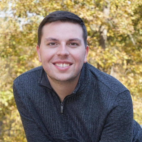 A smiling man with short brown hair, wearing a dark grey sweater, leaning forward slightly, with a backdrop of autumn trees. A smiling man with short brown hair, wearing a dark grey sweater, leaning forward slightly, with a backdrop of autumn trees.