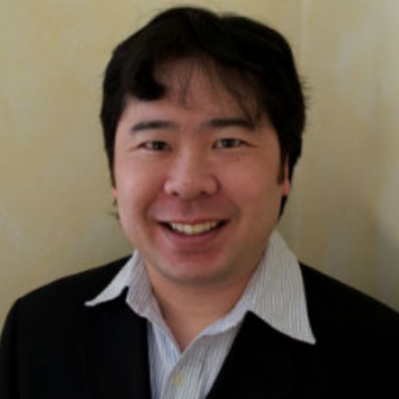 Smiling man with short black hair, wearing a black blazer and a white striped shirt, against a neutral wall background. Smiling man with short black hair, wearing a black blazer and a white striped shirt, against a neutral wall background.
