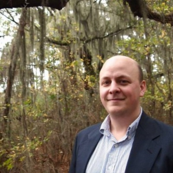 A bald man smiling slightly wearing a striped button up shirt underneath an open navy blue blazer, in front of a forest. A bald man smiling slightly wearing a striped button up shirt underneath an open navy blue blazer, in front of a forest.