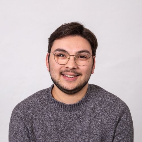 Smiling man with short brown hair, wearing glasses and a dark grey sweater, against a solid off-white background. Smiling man with short brown hair, wearing glasses and a dark grey sweater, against a solid off-white background.