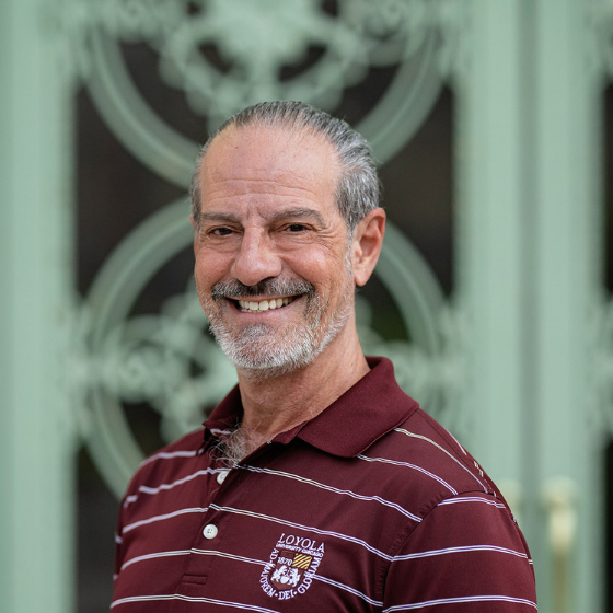 Smiling man with grey hair pulled back into a ponytail, wearing a striped maroon shirt with the Loyola logo on it, pictured outside in front of an ornate gate. Smiling man with grey hair pulled back into a ponytail, wearing a striped maroon shirt with the Loyola logo on it, pictured outside in front of an ornate gate.