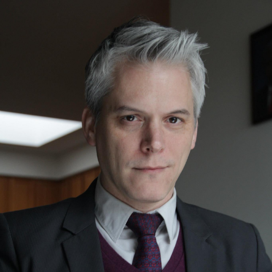 Slightly smiling man with short white hair, wearing a dark grey suit and burgundy tie over a white collared shirt, in front of a classroom background. Slightly smiling man with short white hair, wearing a dark grey suit and burgundy tie over a white collared shirt, in front of a classroom background.
