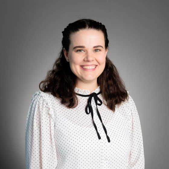 Smiling woman with shoulder-length curly hair, wearing a white top with a black bow at the collar, against a solid grey background. Smiling woman with shoulder-length curly hair, wearing a white top with a black bow at the collar, against a solid grey background.