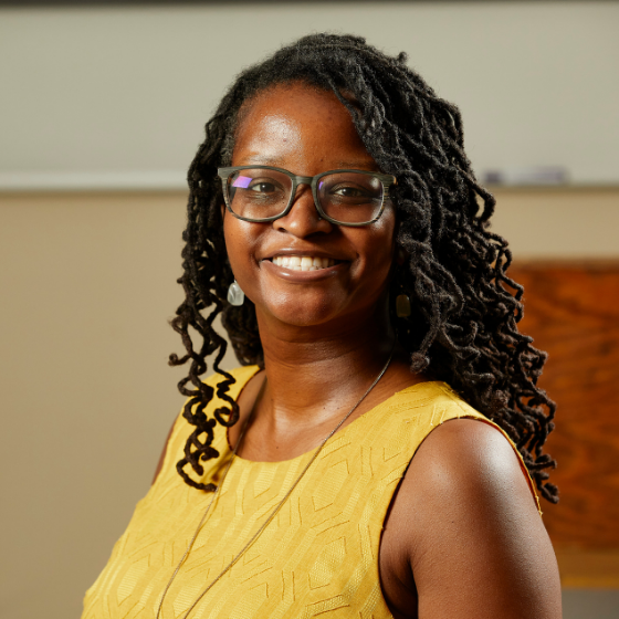 Smiling woman with long curly hair wearing glasses and a yellow top, in front of a school background. Smiling woman with long curly hair wearing glasses and a yellow top, in front of a school background.