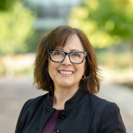 Square headshot of a brunette white woman with glasses, with a college campus in the background Square headshot of a brunette white woman with glasses, with a college campus in the background