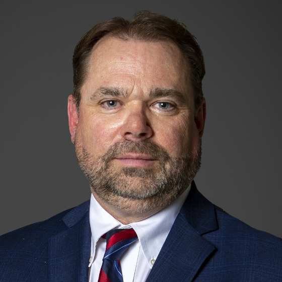 Man with short brown hair smiling faintly and wearing a dark blue suit jacket, white top, and a red tie, in front of a solid dark grey background. Man with short brown hair smiling faintly and wearing a dark blue suit jacket, white top, and a red tie, in front of a solid dark grey background.