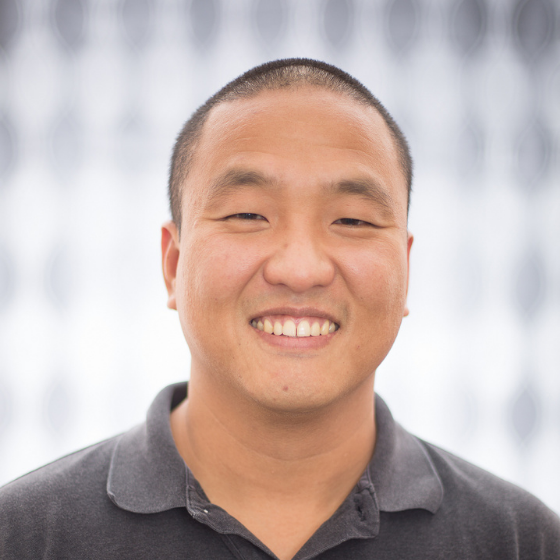 Smiling man with a shaved head, wearing a dark grey shirt with a collar, in front of a checkered curtain. Smiling man with a shaved head, wearing a dark grey shirt with a collar, in front of a checkered curtain.
