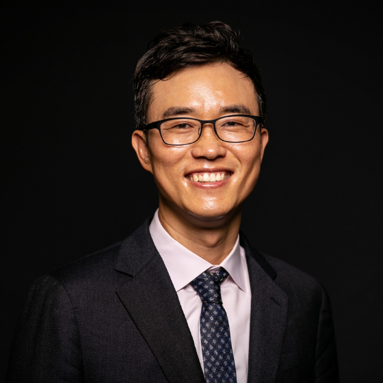 Smiling man with short dark brown hair, wearing glasses, a black suit jacket, and a white top with a dark grey tie, against a solid black background. Smiling man with short dark brown hair, wearing glasses, a black suit jacket, and a white top with a dark grey tie, against a solid black background.