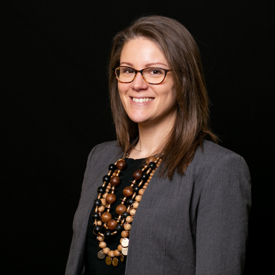 Woman with medium brown hair, smiling and wearing glasses, a dark grey suit jacket, a black top, and several chunky necklaces, against a solid black background. Woman with medium brown hair, smiling and wearing glasses, a dark grey suit jacket, a black top, and several chunky necklaces, against a solid black background.