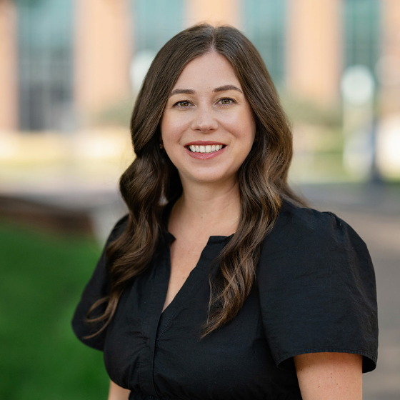Woman smiling with long, wavy brown hair, wearing a black blouse, standing in front of a Loyola University Chicago campus building. Woman smiling with long, wavy brown hair, wearing a black blouse, standing in front of a Loyola University Chicago campus building.