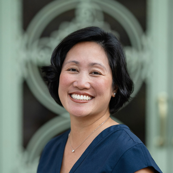 Smiling woman with her black hair in a short bob, wearing a navy blue top, standing in front of a building gate. Smiling woman with her black hair in a short bob, wearing a navy blue top, standing in front of a building gate.