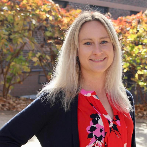 Smiling woman with shoulder length blonde hair, wearing a dark grey cardigan over a floral red top, with greenery in the background. Smiling woman with shoulder length blonde hair, wearing a dark grey cardigan over a floral red top, with greenery in the background.