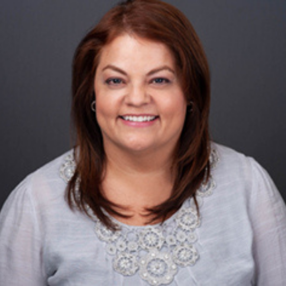 A woman smiling with long brown hair, wearing a light grey blouse, against a solid dark grey background. A woman smiling with long brown hair, wearing a light grey blouse, against a solid dark grey background.