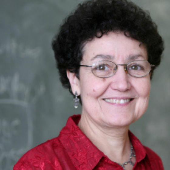 Smiling woman with short, curly dark brown hair, wearing glasses and a collared red top, with a chalkboard in the background. Smiling woman with short, curly dark brown hair, wearing glasses and a collared red top, with a chalkboard in the background.