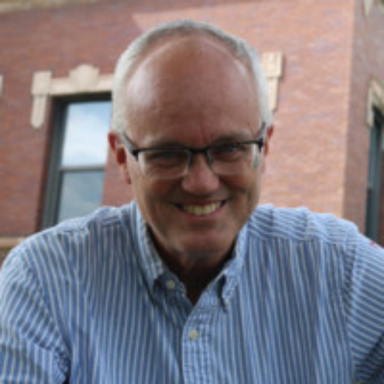 Bald man smiling, wearing glasses, and a light blue button-up top, with a building in the background. Bald man smiling, wearing glasses, and a light blue button-up top, with a building in the background.