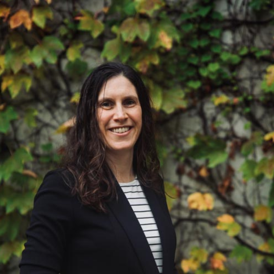 Woman smiling with long wavy brown hair, wearing a black blazer over a white striped top, with greenery in the background. Woman smiling with long wavy brown hair, wearing a black blazer over a white striped top, with greenery in the background.