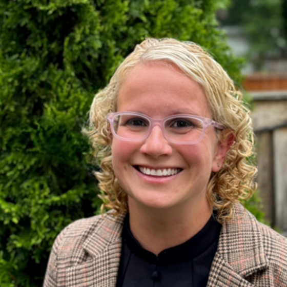 Woman smiling with curly blonde hair, wearing glasses, a gingham tan blazer over a black top, with greenery in the background. Woman smiling with curly blonde hair, wearing glasses, a gingham tan blazer over a black top, with greenery in the background.