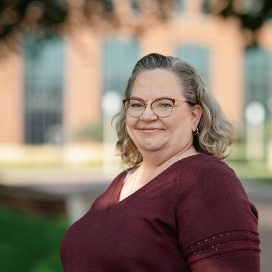 Smiling woman wearing glasses and a maroon colored top, pictured outside in front of a campus building. Smiling woman wearing glasses and a maroon colored top, pictured outside in front of a campus building.