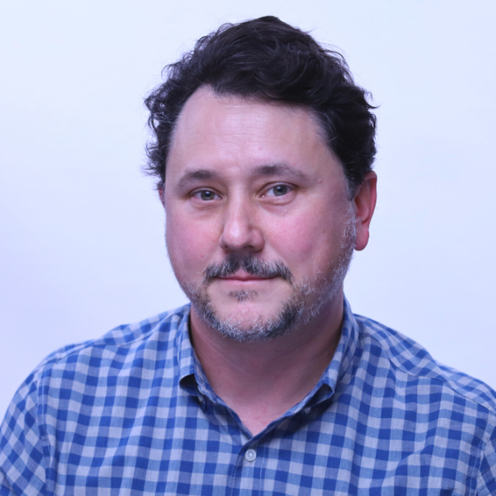A man with short, dark brown hair, smiling slightly, wearing a checkered blue button-up top, against a solid light grey background. A man with short, dark brown hair, smiling slightly, wearing a checkered blue button-up top, against a solid light grey background.