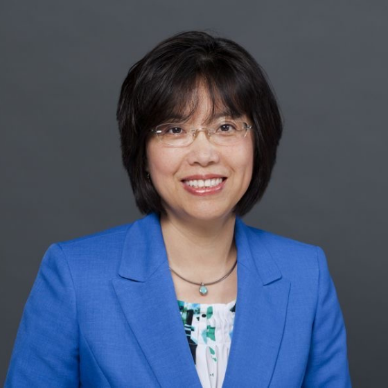 A woman smiling with short black hair, wearing glasses, a necklace, and a blue blazer over a floral light blue top, against a solid dark grey background. A woman smiling with short black hair, wearing glasses, a necklace, and a blue blazer over a floral light blue top, against a solid dark grey background.