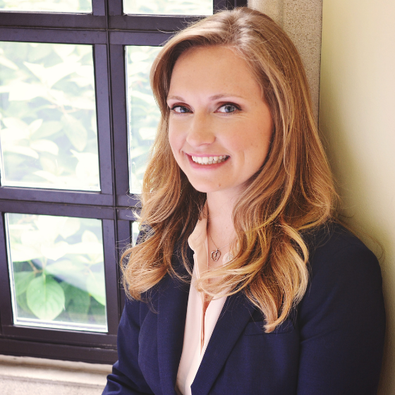 A woman smiling with long, wavy blonde hair, wearing a dark blue blazer over a light pink top, leaning against a windowsill. A woman smiling with long, wavy blonde hair, wearing a dark blue blazer over a light pink top, leaning against a windowsill.