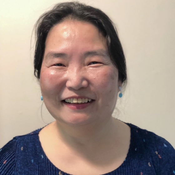 A woman smiling with short black hair, wearing a dark blue top, against an off-white background. A woman smiling with short black hair, wearing a dark blue top, against an off-white background.