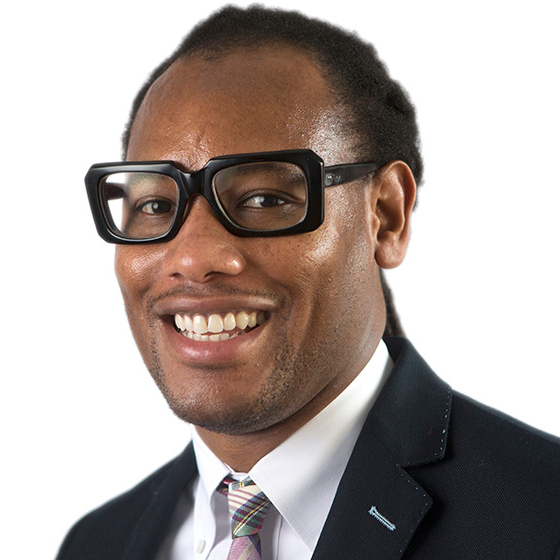 Smiling man with black hair pulled back into locs, wearing thick-rimmed black glasses, a suit jacket, and plaid patterned tie, against a solid white background. Smiling man with black hair pulled back into locs, wearing thick-rimmed black glasses, a suit jacket, and plaid patterned tie, against a solid white background.