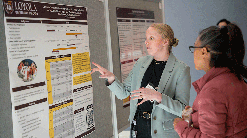 A student points at a research poster and explains it to a woman looking on A student points at a research poster and explains it to a woman looking on