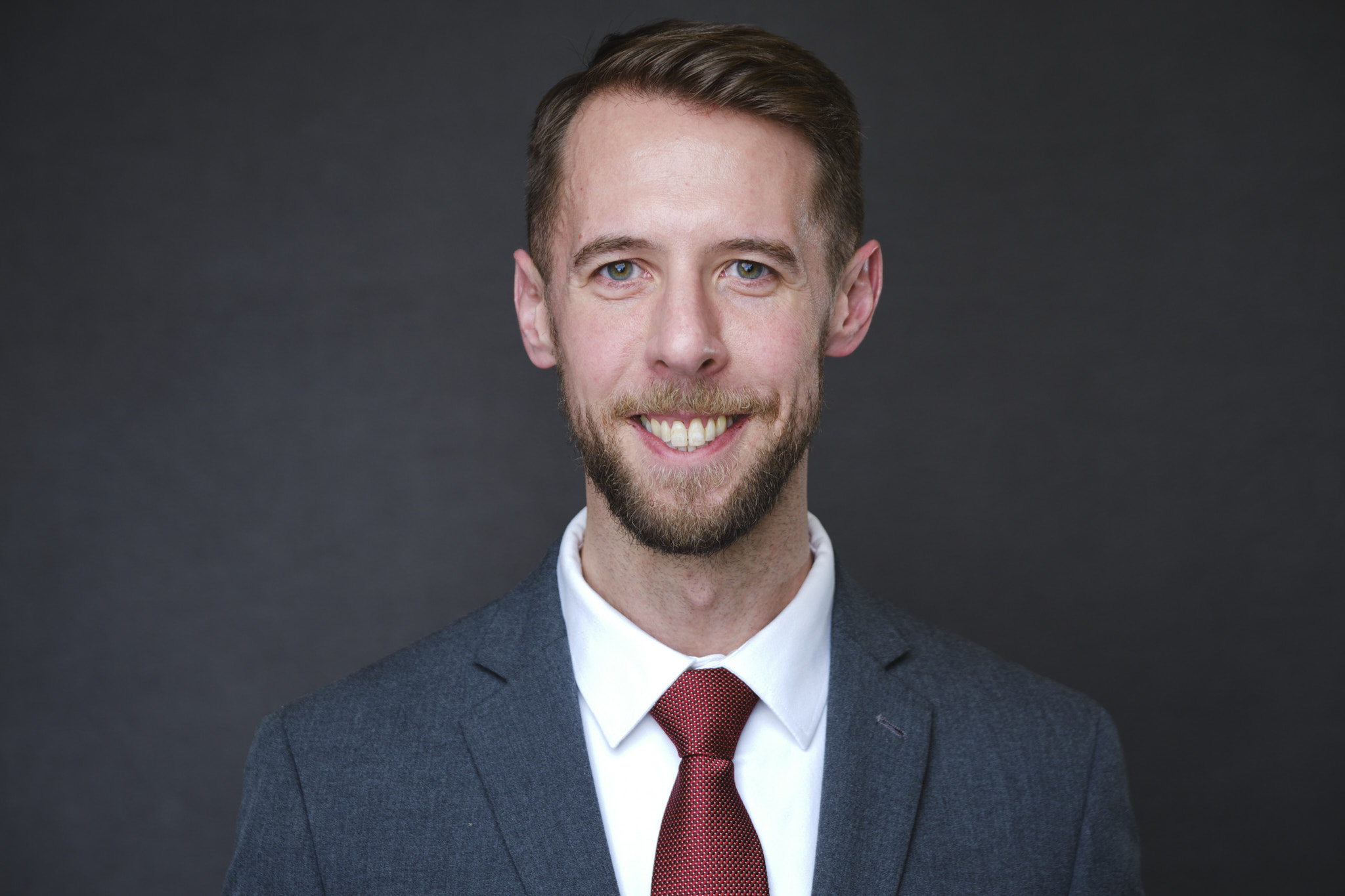 Headshot of a man in a suit and red tie smiling, with a dark background Headshot of a man in a suit and red tie smiling, with a dark background