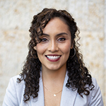 Veronica Arreola smiling, wearing a light gray blazer over a dark top, with shoulder-length dark curly hair, standing against a softly textured neutral background. Veronica Arreola smiling, wearing a light gray blazer over a dark top, with shoulder-length dark curly hair, standing against a softly textured neutral background.