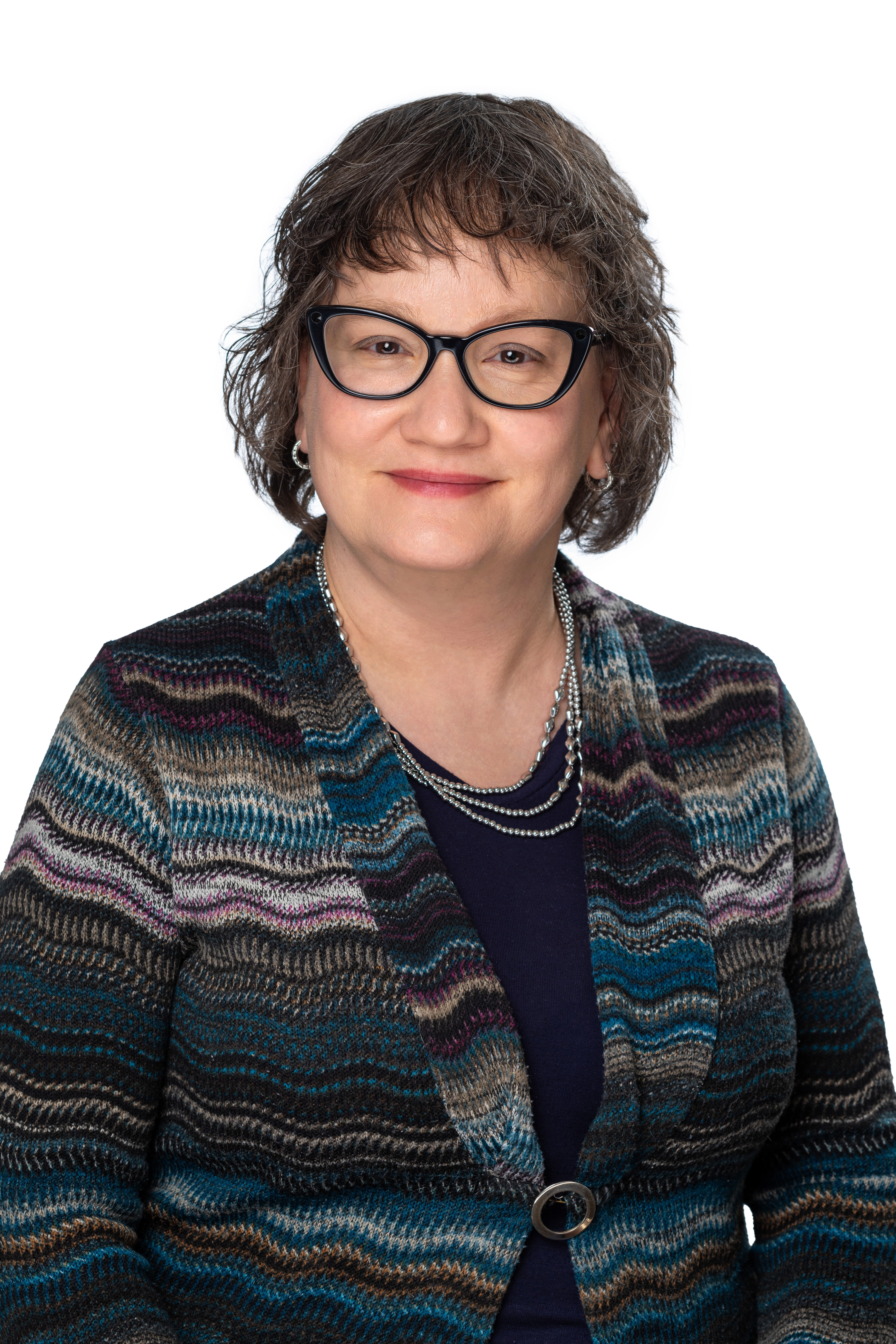 Headshot of a School of Social Work faculty member with short wavy brown hair and dark-framed glasses, wearing a multicolored patterned cardigan over a navy top and layered necklaces, smiling against a light gray background. Headshot of a School of Social Work faculty member with short wavy brown hair and dark-framed glasses, wearing a multicolored patterned cardigan over a navy top and layered necklaces, smiling against a light gray background.