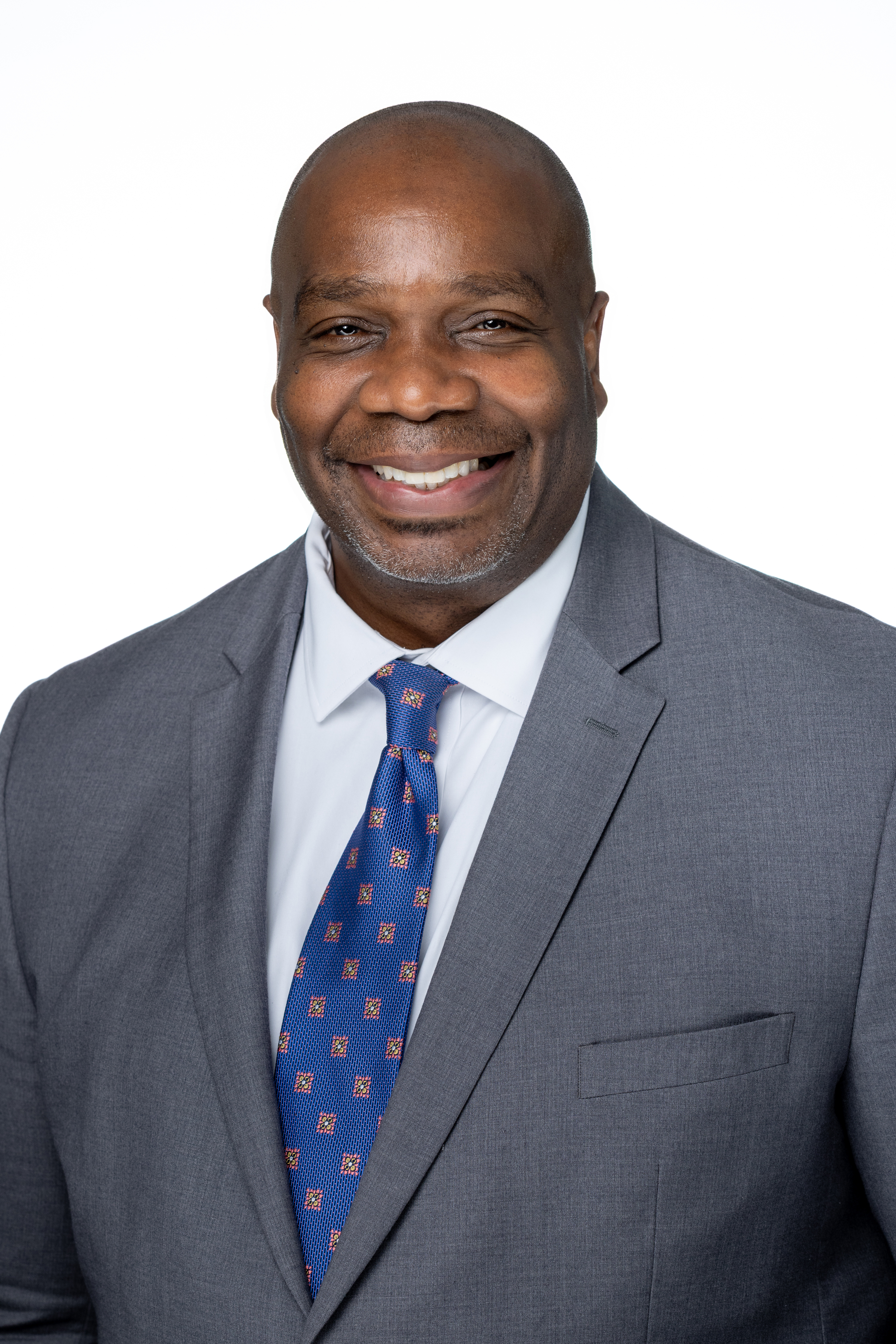 Amzie Moore II, a man with a shaved head and neatly trimmed facial hair, wearing a gray suit jacket, white dress shirt, and a blue patterned tie, smiling against a light gray background. Amzie Moore II, a man with a shaved head and neatly trimmed facial hair, wearing a gray suit jacket, white dress shirt, and a blue patterned tie, smiling against a light gray background.