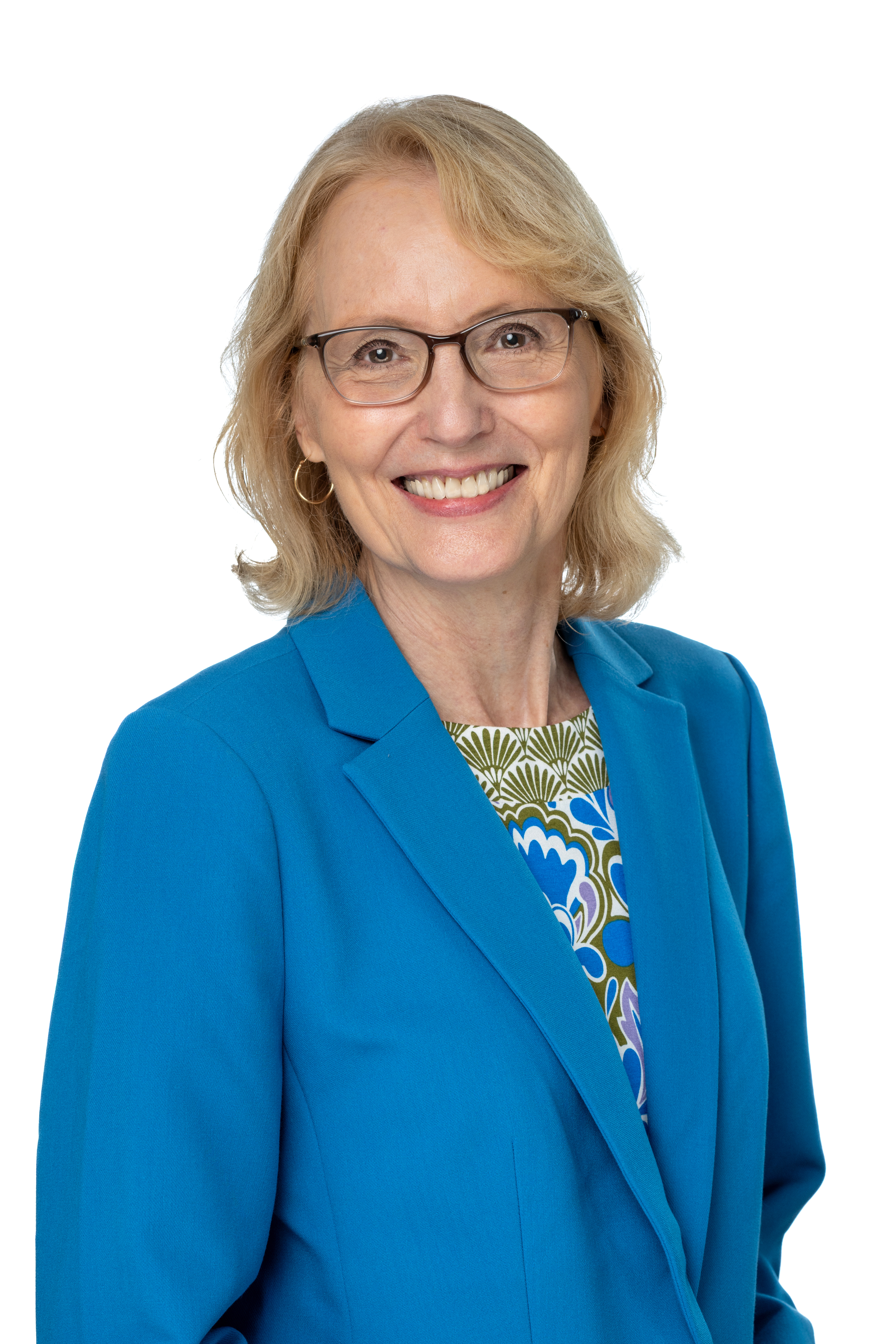 Katherine Tyson McCrea, PhD, smiling and wearing glasses, a bright blue blazer, and a patterned blouse with blue and green accents, against a light gray background. Katherine Tyson McCrea, PhD, smiling and wearing glasses, a bright blue blazer, and a patterned blouse with blue and green accents, against a light gray background.