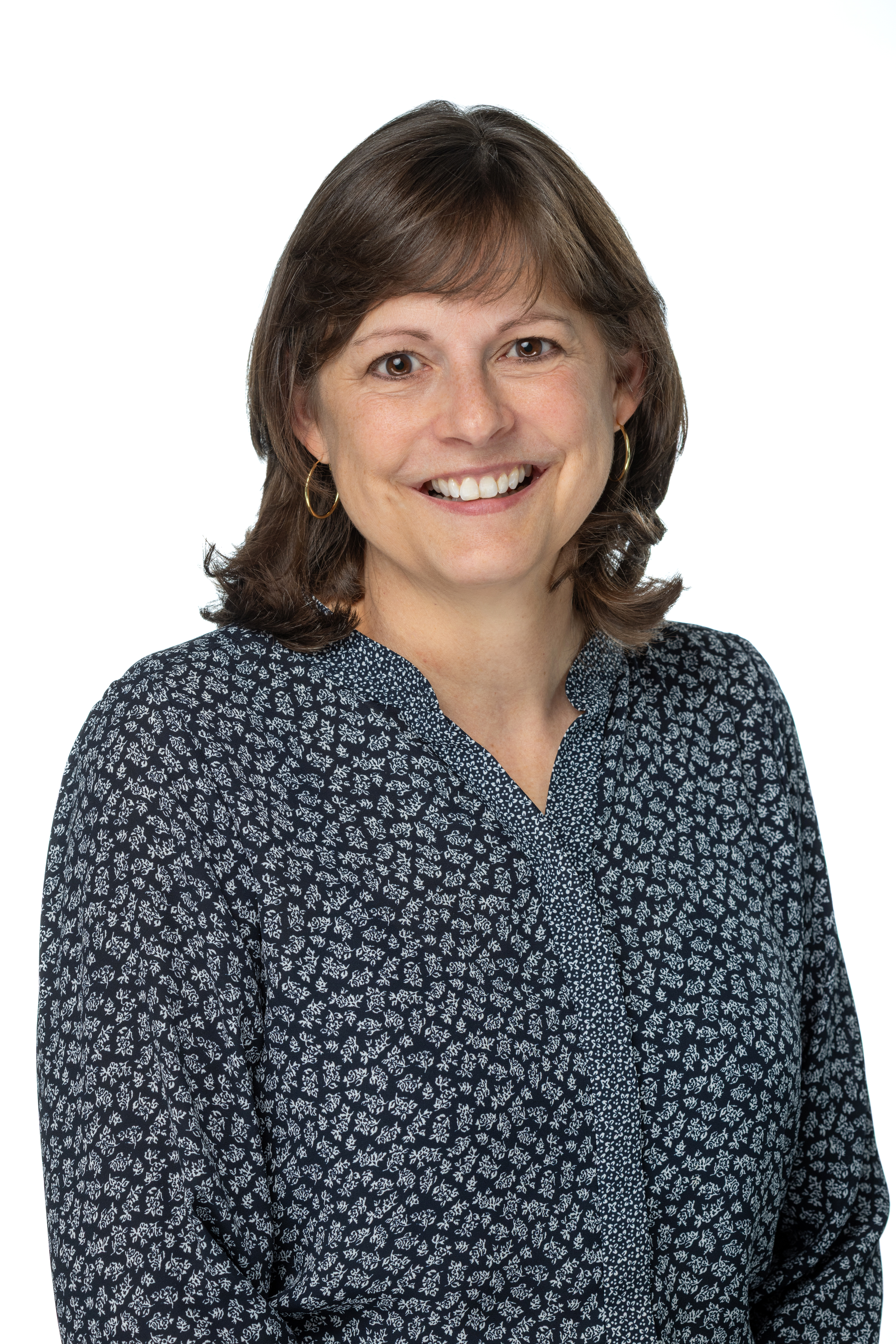 Maria V. Wathen, PhD, smiling and wearing a black top with a textured black-and-white jacket, gold hoop earrings, and a delicate gold necklace, against a white background. Maria V. Wathen, PhD, smiling and wearing a black top with a textured black-and-white jacket, gold hoop earrings, and a delicate gold necklace, against a white background.