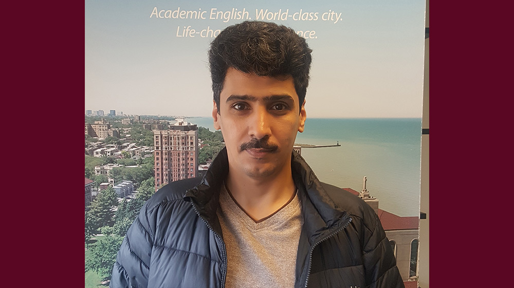 Ahmed wearing a winter jacket standing in front of a ESL Program banner Ahmed wearing a winter jacket standing in front of a ESL Program banner
