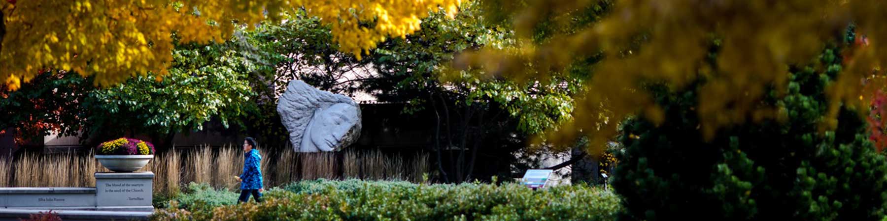 A student walks past a statue in a forested walking path on Loyola University Chicago's Lake Shore Campus A student walks past a statue in a forested walking path on Loyola University Chicago's Lake Shore Campus