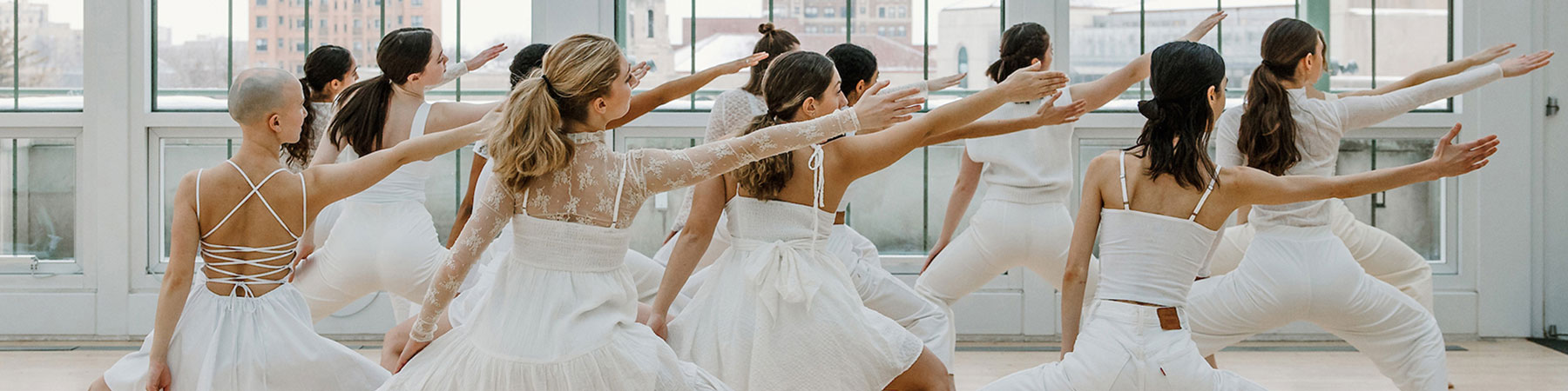 A small group of dancers, facing away from the camera, in various white, dance clothes, move in sync with one another as they perform a dance.