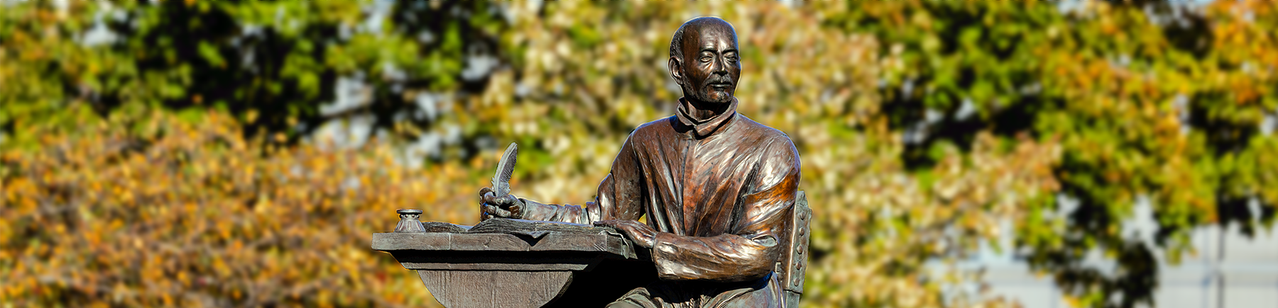 A statue of St. Ignatius sitting and writing at a desk on Lake Shore Campus with fall foliage behind him