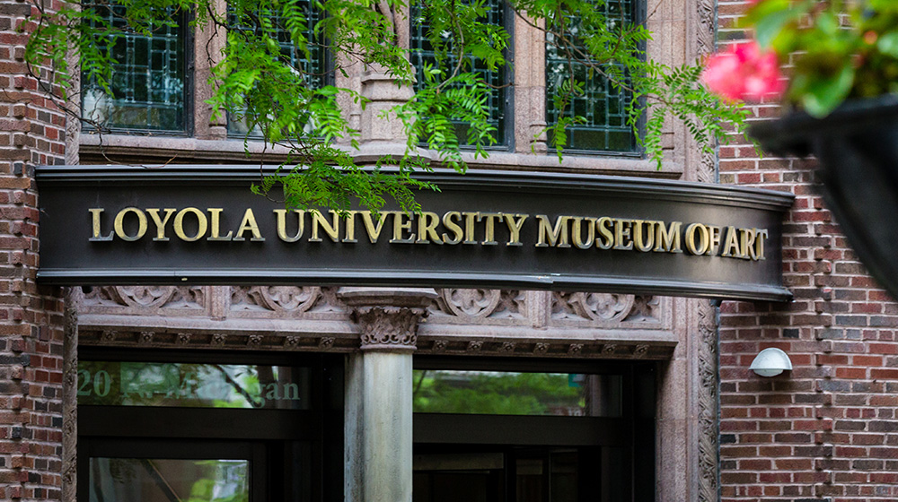 A group of brass serif letters spell out Loyola University Museum of Art on a dark brown background above the entrance to the museum.