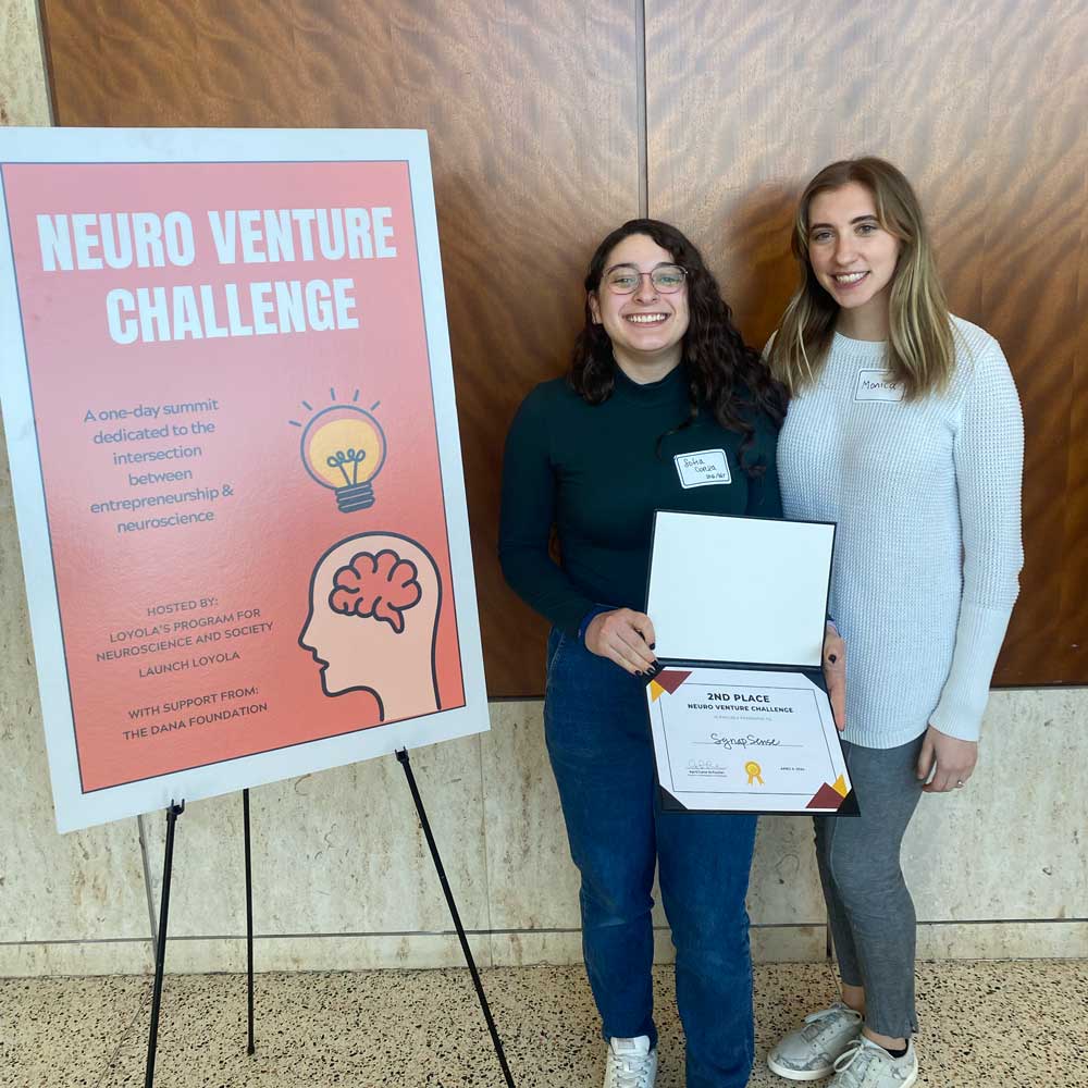 two student posing next to the NeuroVenture Challenge poster holding a second place prize